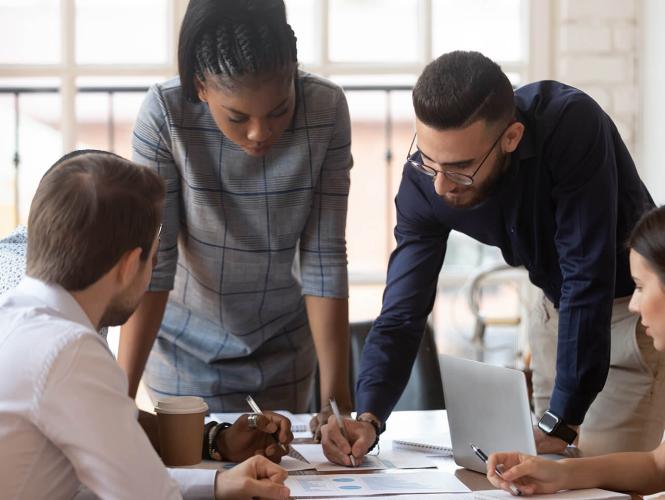 A group of people sitting and standing around a table look through paperwork and documents